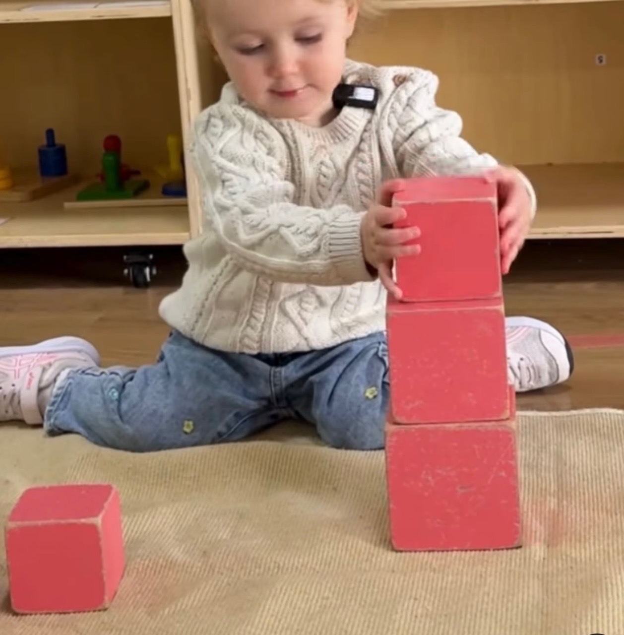 A toddler stacks pink cubes into a tower on the work rug