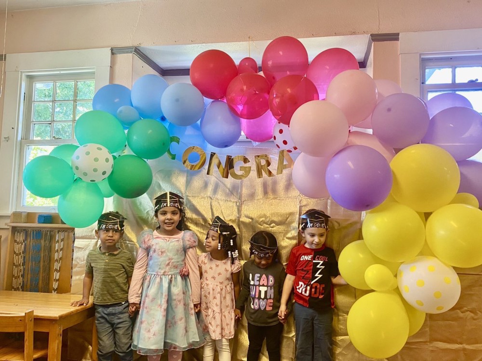 Graduates in decorated caps beneath the rainbow balloon arch