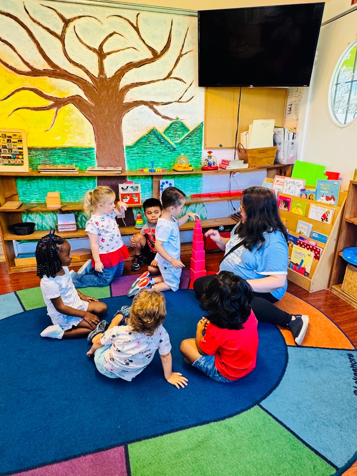 A teacher presents the pink tower to a mixed-age circle beneath the tree mural