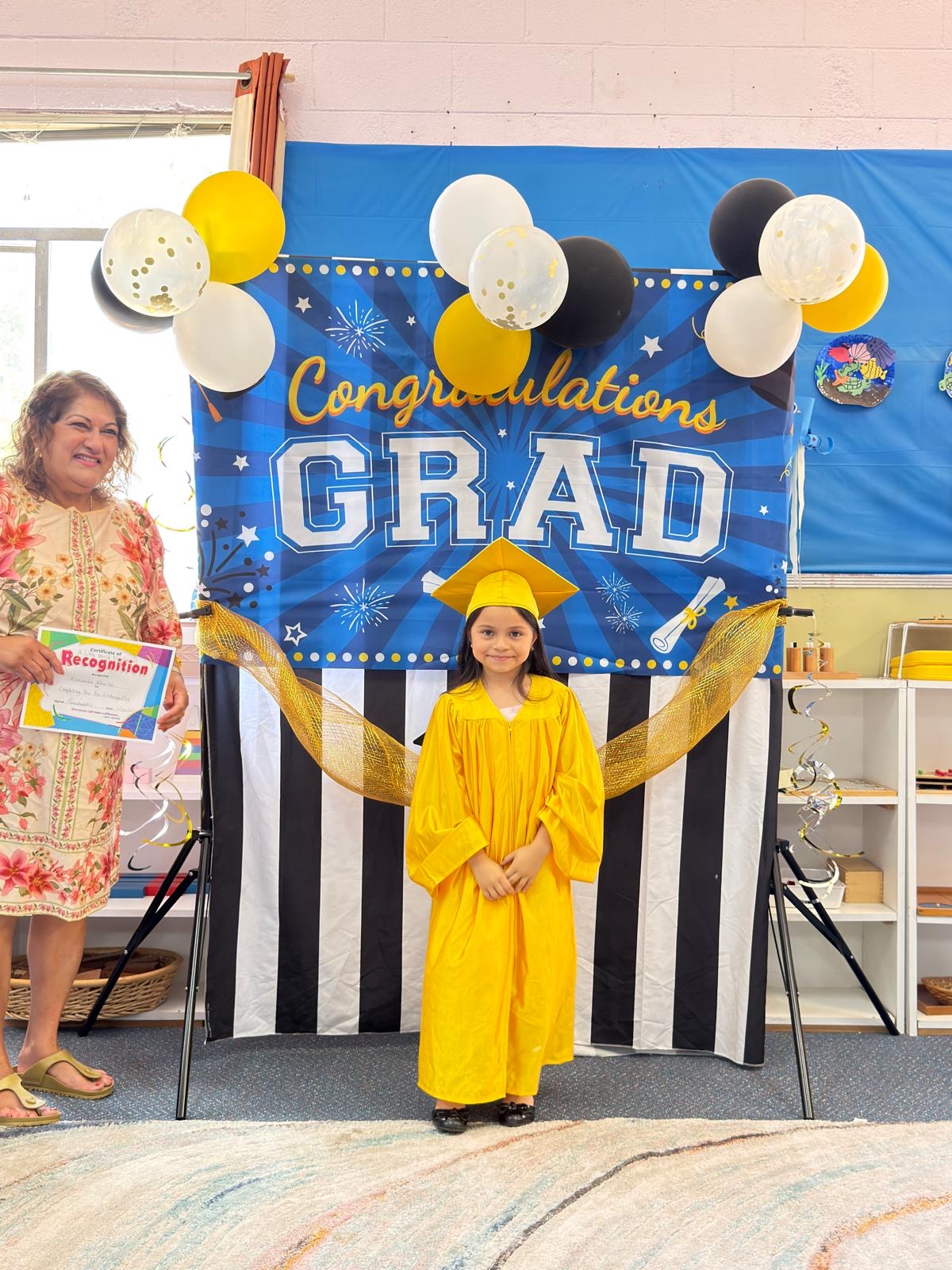 A graduate in a golden gown stands before the Congratulations Grad backdrop