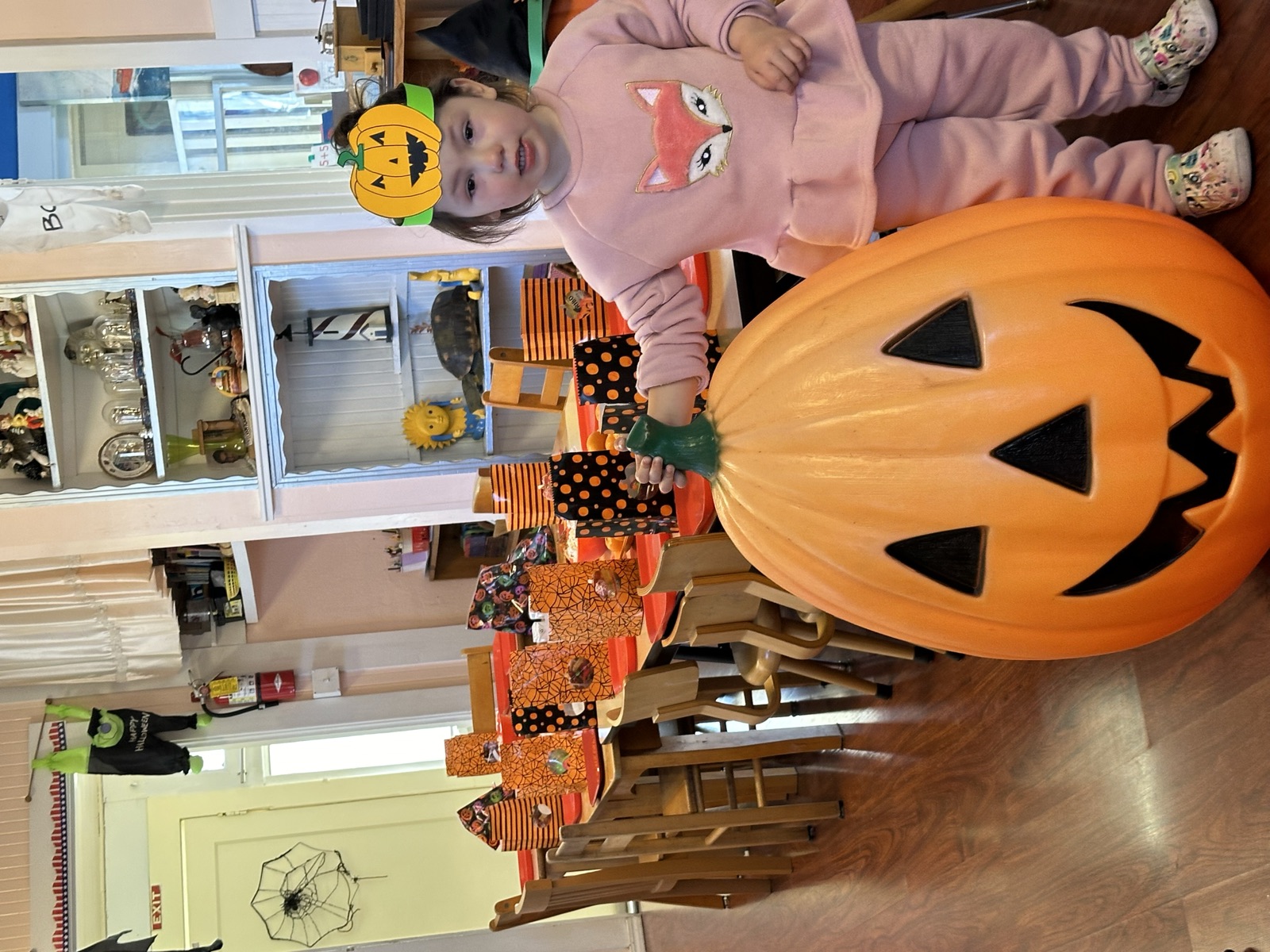 A girl in a pumpkin crown stands beside a giant jack-o'-lantern at the class party table