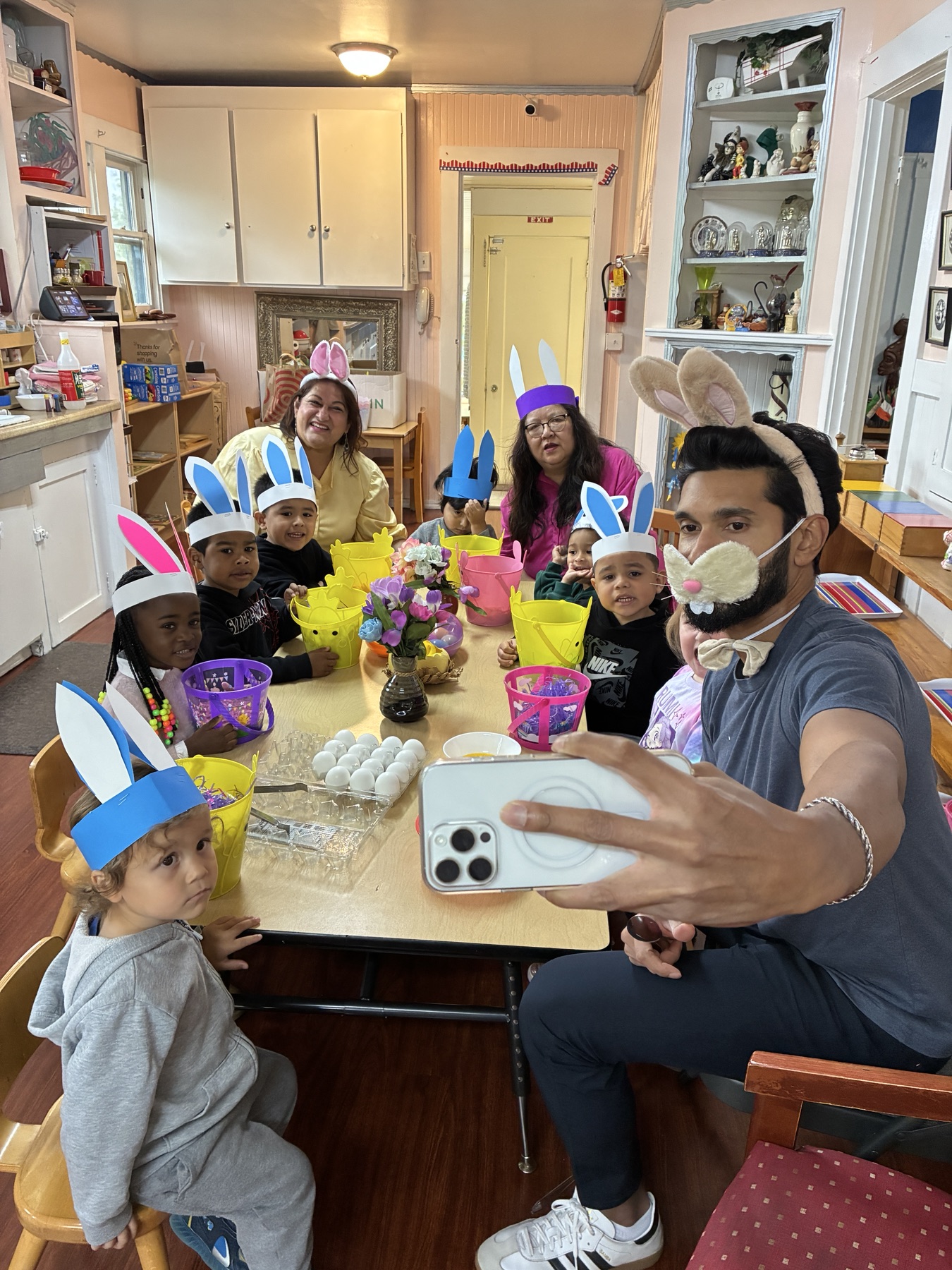 Children in handmade bunny-ear crowns dyeing eggs with teachers and a parent at the spring celebration
